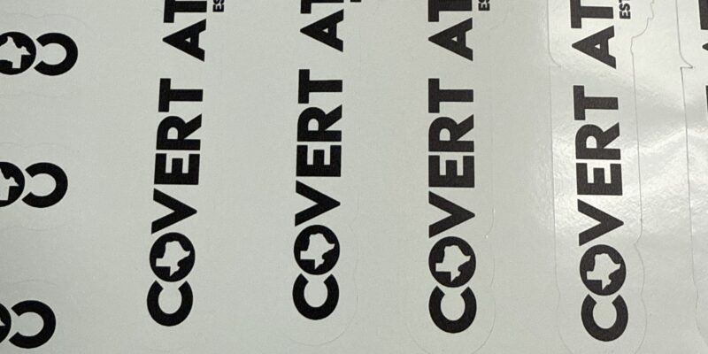 Sheet of custom die-cut vinyl stickers featuring black company branding and logos with precision contour cutting.