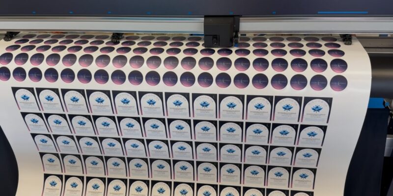 Wide-format digital printer producing bulk custom die-cut vinyl stickers on a roll, featuring circular galaxy-themed logos and arched Blue Lotus Tea labels.